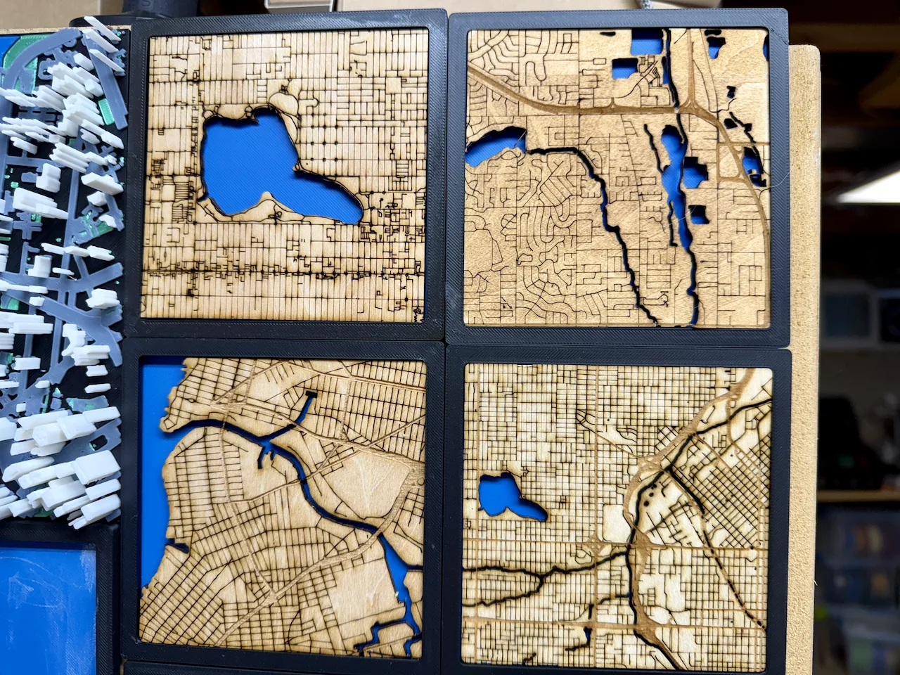 Four framed wooden maps stacked on a workshop shelf beside a printed model